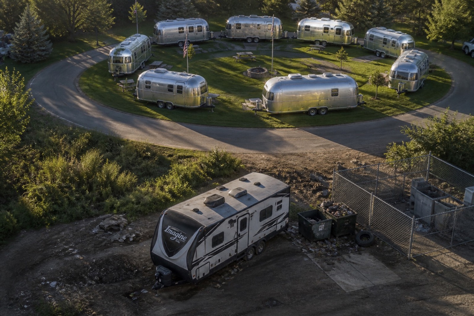 Aerial view of a horseshoe of shiny Airstreams on a pristine loop, with a lone white trailer parked by dumpsters across the road.